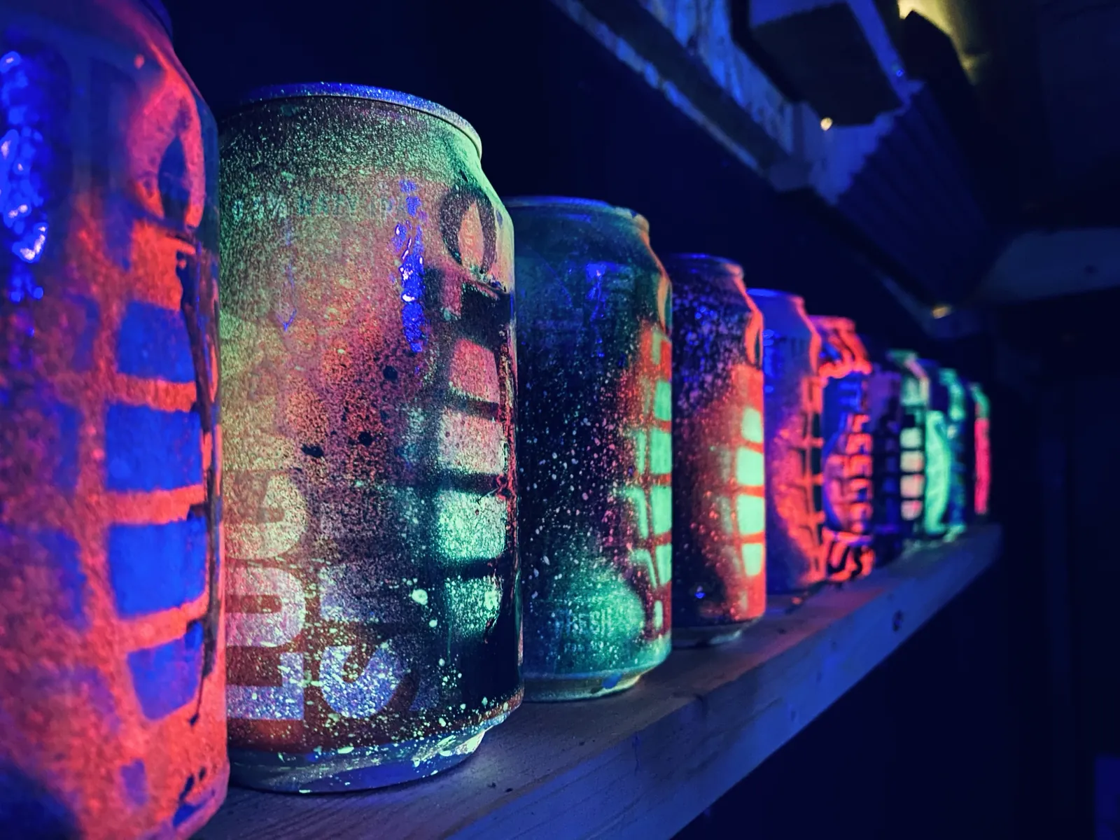 A long shelf of Mangrenades under blacklight, each can glowing a different colour